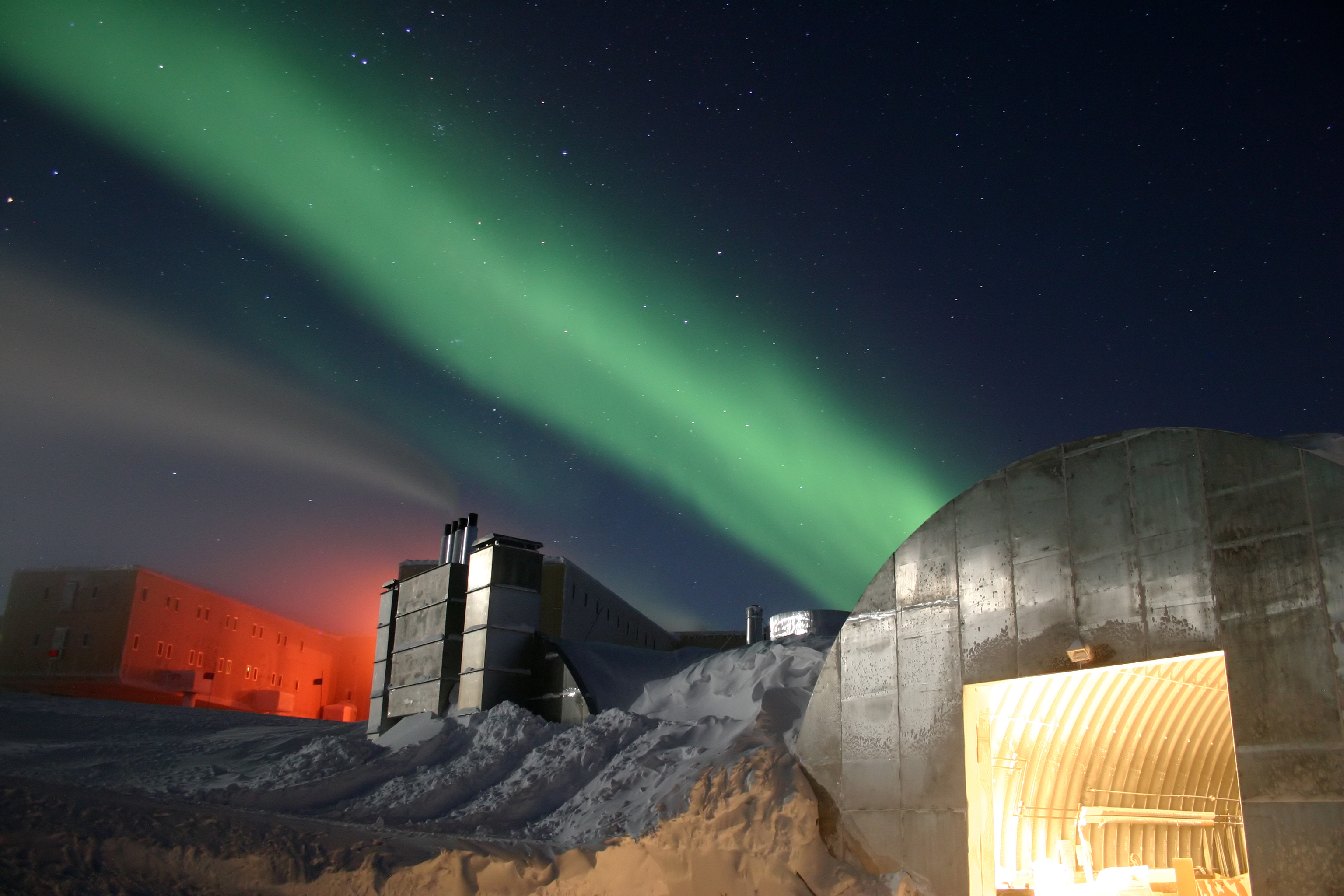 A full moon and 25 second exposure allowed sufficient light into this photo taken at Amundsen-Scott South Pole Station during the long Antarctic night. The station appears at the far left, the power plant in the center, and the mechanic's garage in the lower right. Red lights are used outside during the winter darkness as their spectrum does not pollute the sky, allowing scientists to conduct astrophysical studies without artificial light interference. The background of green light is the aurora australis, which dances through the sky virtually all the time during the long Antarctic night (winter). The photo's surreal appearance makes the station look like a futuristic Mars Station. Image courtesy of the National Science Foundation.