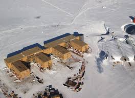 Buildings at the Amundsen-Scott South Pole Station have to be built on stilts at the South Pole to prevent snow buildup. Construction of the latest station began in 1999;  dedication took place in 2008. The ceremonial South Pole and flags can be seen in the background, slightly to the left of center, below the tracks behind the buildings. The actual geographic South Pole is a few more meters to the left, next to the solitary flag. This photo dates to about 2005 and the close of construction, but before the buildings were clad in a final gray sheathing. The older domed station (now dismantled) is partially visible on the right. Image courtesy of the National Science Foundation/ Bill Henriksen.