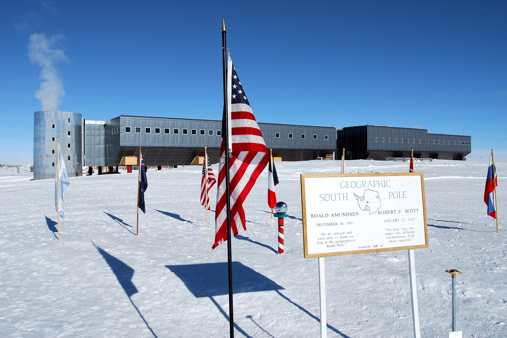 The Amundsen-Scott South Pole Station in the background, the ceremonial South Pole (candy stripe) in the center, and the actual geographic South Pole in the right foreground. Image courtesy of the National Science Foundation.