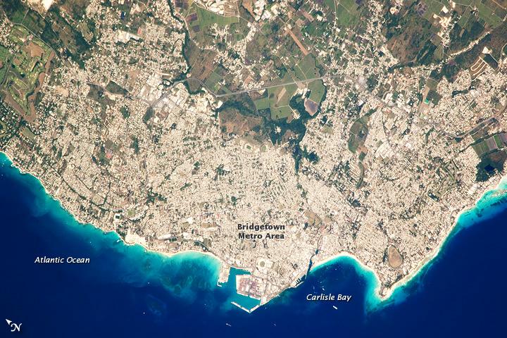 Bridgetown is the capital city of the island nation of Barbados, located to the east of the Lesser Antilles Island chain. While Barbados is considered part of the Lesser Antilles, it is located within the western Atlantic Ocean rather than the Caribbean Sea. Besides being the seat of government, Bridgetown is the largest city in Barbados. It and the surrounding towns that make up the Greater Bridgetown area are located along the southwestern coastline of the island. The metropolitan area is readily recognizable in this astronaut photograph due to the gray and white rooftops and street grids (image center) that contrast with green vegetated fields and riverside areas of the island's interior to the northeast (image top center). Bridgetown is a major port destination for both commercial and cruise ships serving the eastern Caribbean - several ships are visible within Carlisle Bay. Water color in the image changes from light blue along the coastline - indicating shallow water - to the dark blue of deeper water away from the island. Photo courtesy of NASA.