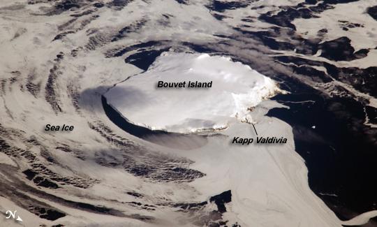 Bouvet Island is one of the most remote islands in the world; Antarctica, over 1,600 km (1,000 mi) to the south, is the nearest land mass. Located near the junction of three tectonic plates, the island is mostly formed from a shield volcano that is almost entirely covered by glaciers. The prominent Kapp (Cape) Valdivia on the northern coastline is a peninsula formed by a lava dome. It is only along the steep cliffs of the coastline that the underlying dark volcanic rock is visible against the white snow and ice blanketing the island. Photo courtesy of NASA.