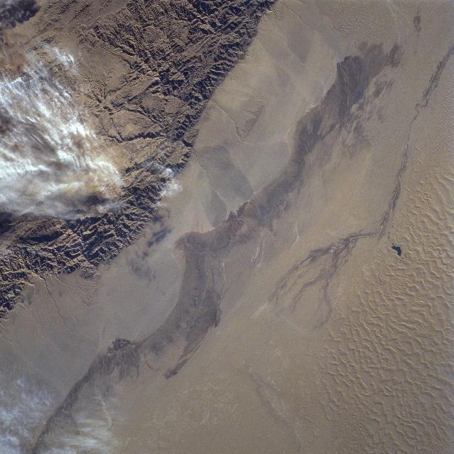 The rugged texture of the Altun Mountains (upper left) that forms part of the northern boundary of the Tibetan Plateau contrasts dramatically with the Takla Makan Desert of western China. Immediately north (to the right) of the mountains are large alluvial fans, gradually sloping downward in elevation. Sand dunes and sand ridges are visible along the northern (lower right) margin of the image. Click on photo for higher resolution. Image courtesy of NASA. 