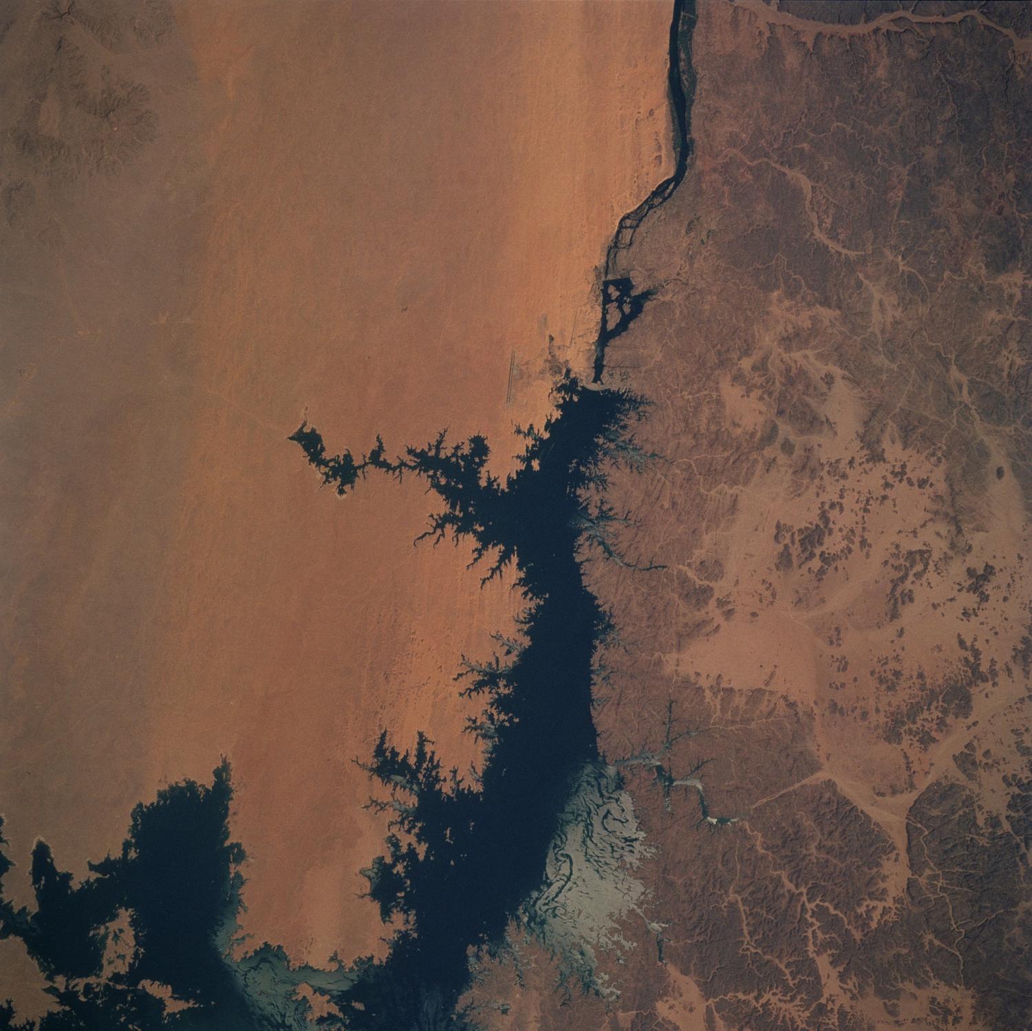 Aswan Dam (upper center of image) and Lake Nasser along the Nile River. The dam controls the flow of the Nile River forming Lake Nasser. The light-colored areas in the lake are where the sun is reflecting off the surface of the water. These areas are fairly calm and not disturbed by wind gusts, enabling the sun glint to show water current patterns on the surface. The Aswan runway is seen as a dark set of lines just west of the Aswan Dam. Click on photo for higher resolution. Image courtesy of NASA. 