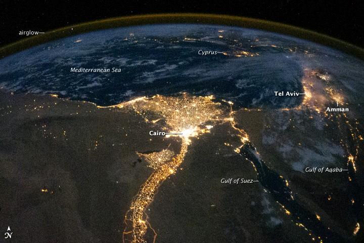 One of the fascinating aspects of viewing Earth at night is how well the lights show the distribution of people. In this view of Egypt, we see a population almost completely concentrated along the Nile Valley, just a small percentage of the country's land area. 
The Nile River and its delta look like a brilliant, long-stemmed flower in this astronaut photograph of the southeastern Mediterranean Sea. The Cairo metropolitan area forms a particularly bright base of the flower. The smaller cities and towns within the Nile Delta tend to be hard to see amidst the dense agricultural vegetation during the day. However, these settled areas and the connecting roads between them become clearly visible at night. Likewise, infrastructure and urbanized regions along the Nile River also become apparent.
Another brightly lit region is visible along the eastern coastline of the Mediterranean, the Tel-Aviv metropolitan area in Israel (image right). To the east of Tel-Aviv lies Amman, Jordan. The two major water bodies that define the western and eastern coastlines of the Sinai Peninsula, the Gulf of Suez and the Gulf of Aqaba, are outlined by lights along their coastlines (image lower right). The city lights of Paphos, Limassol, Larnaca, and Nicosia are visible on the island of Cyprus (image top).
Scattered blue-grey clouds cover the Mediterranean Sea and the Sinai, while much of northeastern Africa is cloud-free. The thin yellow-brown band tracing the Earth's curvature at image top is airglow, a faint band of light emission that results from the interaction of atmospheric atoms and molecules with solar radiation at approximately 100 km (60 mi) altitude. Photo courtesy of NASA.