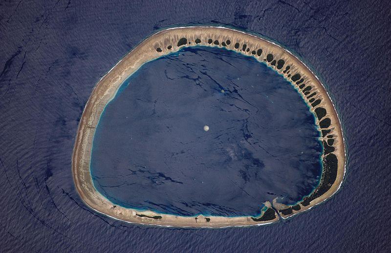 Classically shaped Nukuoro Atoll is one of 607 islands that make up the Federated States of Micronesia. It is also a municipality of the state of Pohnpei, Federated States of Micronesia. The atoll has a population of only several hundred people. The total area, including the lagoon, is 40 sq km, with a land area of only 1.7 sq km, which is divided among more than 40 islets that lie on the northern, eastern, and southern sides of the lagoon. By far the largest islet is Nukuoro islet in the southeast, which is the center of population and the capital of the municipality. Vegetation is most prominent on the northeast and east portions of the atoll that face the dominant Easterly winds.  Swells driven by these winds can be seen approaching from the east-southeast (right) and wrapping around the atoll to produce an interference pattern on the downwind side. Water in the lagoon is calm in comparison. A coral head appears dead center of the photo. Image courtesy of NASA. 