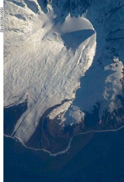 The northeastern slopes of Big Ben volcano on Heard Island are visible in shadow at the top right of this photo. Mawson Peak (small circle in the upper center of the image), sits within a breached caldera (collapsed and empty magma chamber beneath a volcano) on the southwestern side of the volcano - the shadow cast by Mawson Peak points directly to the crescent-shaped caldera rim.  This image, taken during the Southern Hemisphere summer, also shows the dark brown volcanic rock that forms the island. The southwest-pointing tongue of ice is Gotley Glacier. Image credit: NASA.