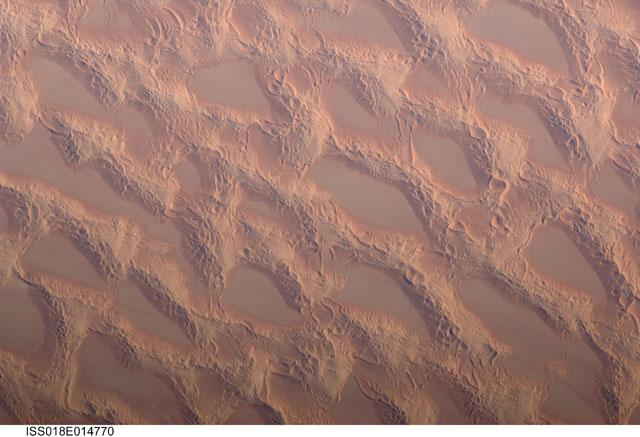 Photo shows classic large and small sand masses of the central Sahara Desert, where wind is a more powerful land-shaping agent than water. "Draa" dunes (from the Arabic for "arm") are very large masses of sand, and they appear here as the broad network of yellow-orange sand masses, with smooth-floored, almost sand-free basins between them. These sand masses lie in the western part of Libya's vast Marzuq Sand Sea. Geologists think that the draa of the Marzuq were probably formed by winds different from the prevailing north-northeast winds of today. Numerous smaller dunes have developed on the backs of the draa. Three distinct dune types are visible: longitudinal dunes, which are more or less parallel with the north winds; transverse dunes, which are usually more curved and formed at right angles to the wind; and star dunes, in which several linear arms converge towards a single peak. Image courtesy of NASA.
