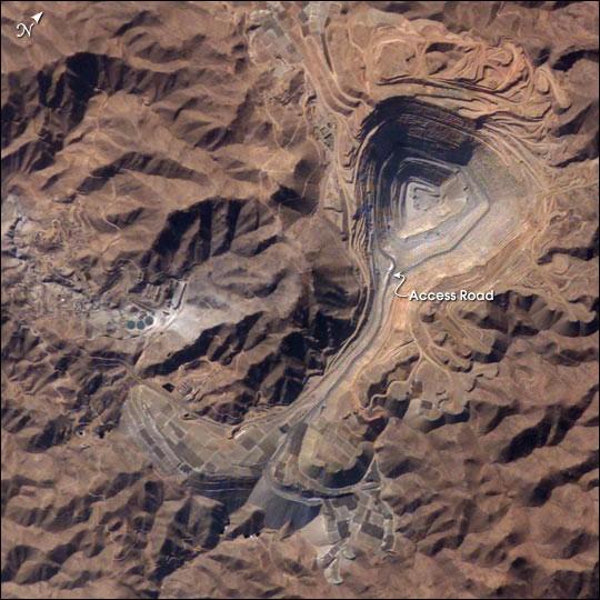 The rugged, mineral-rich Andes support some of the world's biggest mines (gold, silver, copper, and more). This image looks down the bull's-eye of Peru's Toquepala copper mine, a steep sided and stepped open-pit mine. Mid-afternoon sunlight on the arid slopes of the central Andes mountains provides an accent to the mine contours. At the surface, the open pit is 2.5 km (1.5 mi) across, and it descends more than 700 m (2,300 ft) into the earth. A dark line on the wall of the pit is the main access road to the bottom. Spoil dumps of material mined from the pit are arranged in tiers along the northwest lip of the pit. Numerous angular leaching fields appear lower right, and the railroad to the coast is a line that exits the image center left. The railroad was built to export Toquepala's copper and connects the coastal port of Ilo, 95 km (60 mi) to the southwest. Photo courtesy of NASA.