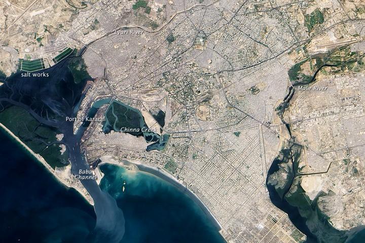 A centuries-old settlement that now ranks among the world's largest, Pakistan's seaport city of Karachi mixes intense urbanization with remnants of a natural environment. This true-color satellite image of Karachi shows the southwestern edges of the city.
Two rivers, the Lyari and Malir, pass through Karachi en route to the Arabian Sea. The Lyari River passes north of the Port of Karachi, and expansive salt works, appearing as large geometric areas of green, line the northern edge of that river's delta. East of the port, the Malir River flows southward toward the sea. 
Between the port and the Arabian Sea, a barrier island runs northwest-southeast (left edge of the image). East of this island and west of the port sits a large expanse of water and mangroves. Water in the Lyari River Delta must empty into the sea through the Baba Channel. Sediment colors the water flowing through that channel a lighter color than the surrounding ocean. Immediately east of the Port of Karachi lie two more mangroves, the larger of which is named Chinna Creek.
Many of Karachi's oldest structures and most popular tourist attractions appear in the city center just northeast of the port, but street grids cover almost all of the available land outside of the mangroves. Photo courtesy of NASA.