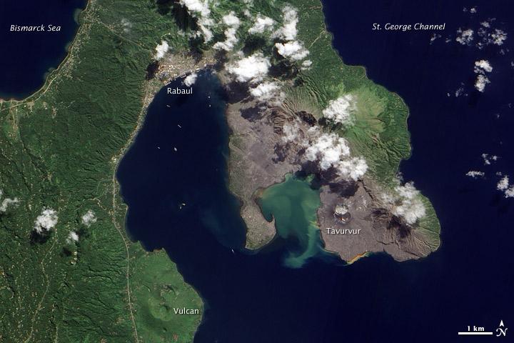 Volcanic cones and settlements mingle along the margins of the Rabaul Caldera on the northeastern tip of Papua New Guinea's New Britain Island. Shaped like a giant cauldron, the caldera is the remaining rim of an ancient volcano. Small volcanic cones, some of which are still active, sit on the rim. Spanning roughly 8 by 14 km (5 by 9 mi), the caldera is flooded by Blanche Bay. The largely submerged caldera long formed a natural harbor for what had been New Britain's largest city, but a major eruption forced the evacuation of Rabaul City in 1994. Although the city ceased to bustle with activity, at least one volcanic cone remained active for years afterward.
The landscape in this satellite image appears as a mixture of vegetated and bare ground. Vegetation almost completely coats the western side of Rabaul Caldera. Volcanic cones line the northeastern margin of the caldera, and around these cones, the land is mostly rocky and barren.
Tavurvur, near the southern end of the exposed caldera rim, was one of the volcanoes to erupt explosively in 1994. The other was Vulcan, on the western side of the caldera. While vegetation has revived on the slopes of Vulcan, rocks predominate at Tavurvur, evidence of more recent eruptive activity.
Remnants of settlements linger along the caldera's northwestern margin and on a promontory near the caldera's center. Prior to the explosive eruption in 1994, the city of Rabaul had been the provincial capital of East New Britain. After the eruption, the capital was moved to the coastal city of Kokopo, some 20 km (12 mi) to the southeast. Photo courtesy of NASA.