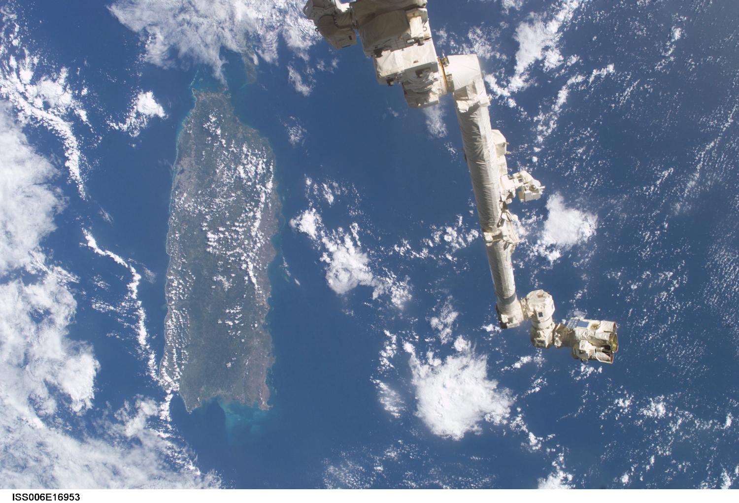 The Space Station Remote Manipulator System (SSRMS) or Canadarm2 appears against the Caribbean Sea in this view taken from the International Space Station. Puerto Rico is the large island on the left side of the frame. Image courtesy of NASA.