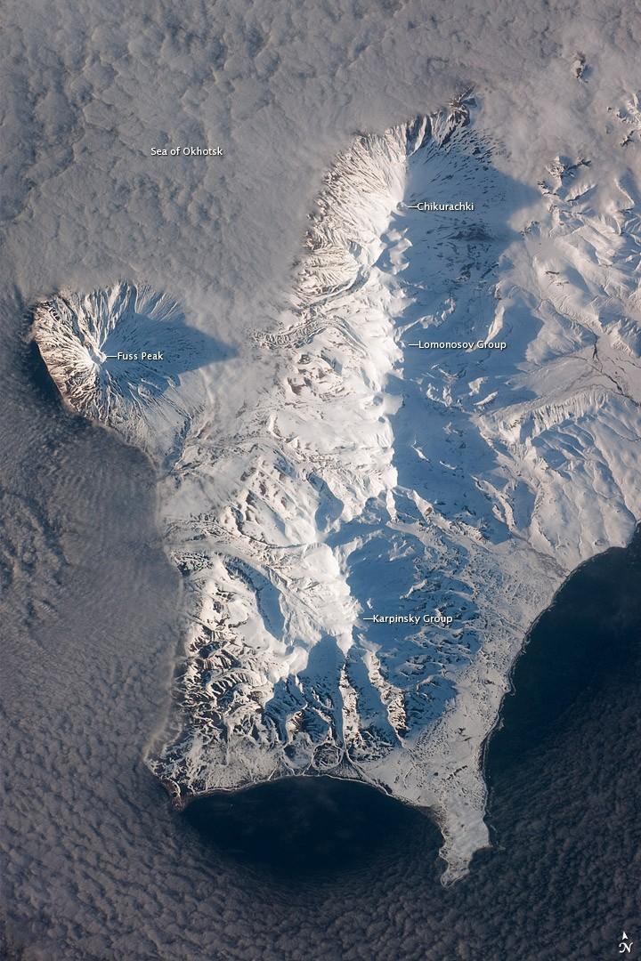 The Kuril Island chain is built from a line of volcanoes, an island arc, that extends from Russia's Kamchatka Peninsula to northern Japan. Island arcs form along an active boundary between two tectonic plates, with one being driven beneath the other (subduction). Magma generated by subduction feeds volcanoes - and eventually volcanic islands - over the subduction boundary. Paramushir Island in the northern Kurils is an example of a large island built by several volcanoes over geologic time. This astronaut photograph shows the southern end of Paramushir Island after a snowfall. The western slopes of the mountains are brightly illuminated, while the eastern slopes are in shadow.
Four major volcanic centers create this part of the island. Fuss Peak (image center left) is an isolated stratovolcano connected to the main island via an isthmus. Fuss Peak last erupted in 1854.
The southern tip of the island is occupied by the Karpinsky Group of three volcanic centers. A minor eruption of ash following an earthquake occurred on this part of the island in 1952. 
The Lomonosov Group to the northeast (image center) includes four cinder cones and a lava dome.
The most recent volcanic activity on Paramushir Island occurred in 2008 at the Chikurachki cone located along the northern coastline of the island at image top center. The summit of this volcano, 1,816 m (5,958 ft) above sea level, is the highest on Paramushir Island.
Much of the Sea of Okhotsk visible in the image is covered with low clouds that often form around the islands in the Kuril chain. The clouds are generated by moisture-laden air passing over the cool sea/ocean water, and they typically wrap around the volcanic islands. Image courtesy of NASA.