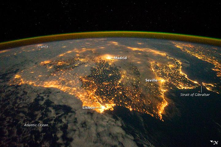 The city lights of Spain and Portugal define the Iberian Peninsula in this photograph taken from the International Space Station (ISS) on 4 December 2011. Several large metropolitan areas are visible, marked by their relatively large and brightly lit areas, such as the capital cities of Madrid, Spain - located near the center of the peninsula's interior - and Lisbon, Portugal - located along the southwestern coastline. The ancient city of Seville, visible at image right to the north of the approximately 14 km (9 mi)-wide Strait of Gibraltar, is one of the largest cities in Spain. The view is looking outwards from the ISS towards the east.

The network of smaller cities and towns in the interior and along the coastline attest to the areal extent of human presence on the Iberian landscape. Blurring of the city lights is caused by thin cloud cover (image left and center), while the cloud tops are dimly illuminated by moonlight. Though obscured, the lights of France are visible near the horizon line at image upper left, while the lights of northern Africa are more clearly discernable at image right. The gold to green line of airglow, caused by excitation of upper atmosphere gas molecules by ultraviolet radiation, parallels the horizon line (or Earth limb). Image courtesy of NASA.
