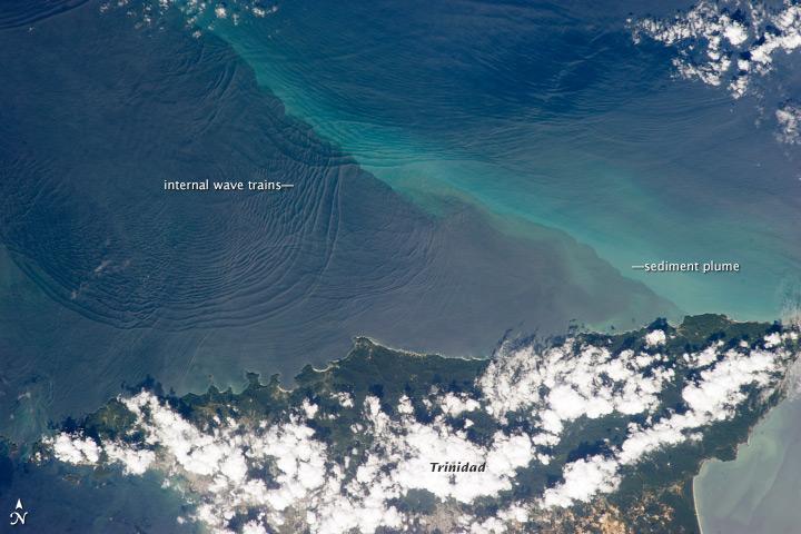 This space station photo of 18 January 2013, shows the north coast of Trinidad and a series of subtle, interacting arcs in the southeastern Caribbean Sea. These are known as "internal waves," the surface manifestation of slow waves that move tens of meters beneath the sea surface. Internal waves produce enough of an effect on the sea surface to be seen from space, but only where they are enhanced due to reflection of sunlight, or sunglint, back towards the space station. 
The image shows at least three sets of internal waves interacting. The most prominent set (image top left) shows a packet of several waves moving from the northwest due to the tidal flow towards the north coast of Trinidad. Two less prominent, younger sets can be seen further out to sea. A very broad set enters the view from the north and northeast, and interacts at image top center with the first set. All the internal waves are probably caused by the shelf break near Tobago (outside the image to top right). The shelf break is the step between shallow seas (around continents and islands) and the deep ocean. It is the line at which tides usually start to generate internal waves. 
Over the island of Trinidad, the heating of the land surface sets off the growth of cumulus clouds. Off the coast, a light blue northwest-southeast trending plume at image center is sediment embedded in the Equatorial Current (also known as the Guyana Current). The current is transporting material to the northwest - in almost the opposite direction of the internal waves. The current flows strongly from east to west around Trinidad, all the way from equatorial Africa, driven by year-round easterly winds. Seafarers in the vicinity of Trinidad are warned that the current - and its local reverse eddies - make navigation complicated and sometimes dangerous for smaller craft in these waters.
Astronauts also have observed internal waves in other parts of the world, such as San Francisco and the Straits of Gibraltar. Photo courtesy of NASA.