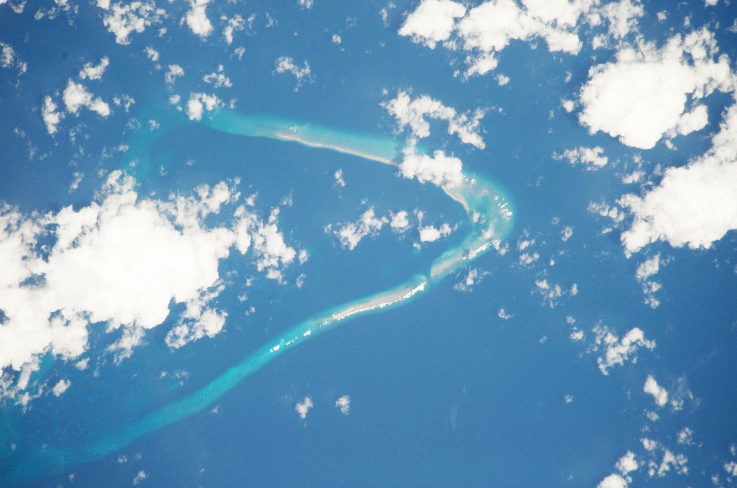Astronaut photo of Kingman Reef, a largely submerged, uninhabited triangular structure in the North Pacific Ocean, roughly halfway between the Hawaiian Islands and American Samoa. The reef lagoon is 82 m (270 ft) deep at the western part and the total enclosed area is about 76 sq km (29 sq mi).  The east-west and northwest-southeast trending small strips of dry land (gray color) are composed of coral rubble and giant clamshells. The highest point on the reef is less than 1.5 m (5 ft) above sea level, which is awash most of the time, making Kingman Reef a maritime hazard. Image courtesy of NASA.