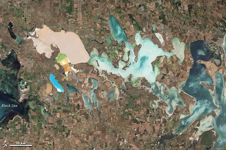 From mainland Ukraine, the Crimean Peninsula extends southward, bordered on the west by the Black Sea and on the east by the Sea of Azov. Stretching across the peninsula is a network of shallow, marshy inlets sprawling over roughly 2,600 sq km (1,000 sq mi). This network of lagoons is known as Syvash (also Sivash or Sivas). During the summer months, the warmed marsh waters give off unpleasant odors, lending the region the nicknames of "Rotten Sea" or "Putrid Sea."
This natural-color satellite image shows the shallow waters and varied chemical composition of the Syvash lagoons contribute to their unearthly colors of peach, mustard, lime green, blue, blue-green, beige, and brown. Thick layers of silt coat the bottoms of the shallow marshes, which are rich enough in mineral salts to supply a local chemical plant.
Surrounding the marshy areas are mostly agricultural fields. Urbanized areas appear along the shores of the Black Sea, and highways curve and zigzag across the peninsula. Outside of the marshes, the land in this area is generally a level plain of arid steppe. In colder months, frosts alternate with thaws, and fogs are frequent. Photo courtesy of NASA.