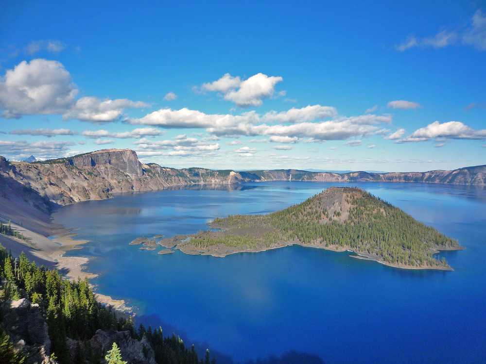 Crater Lake, Oregon in summer I. A view from the West Rim, taken near the Lightning Springs Picnic Area. The island’s name is Wizard Island. 
Formed 7,700 years ago when a violent eruption triggered the collapse of a tall peak, scientists today marvel at the lake’s purity: fed by rain and snow. It is the deepest lake in the US and one of the most pristine on earth. Artists, photographers, and sightseers gaze in wonder at its blue water and stunning setting atop the Cascade Mountain Range. Photo courtesy of the US National Park Service.
