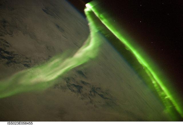 Among the views of Earth afforded crew members aboard the International Space Station (ISS), surely one of the most spectacular is of the aurora. These ever-shifting displays of colored ribbons, curtains, rays, and spots are most visible near the North (aurora borealis) and South (aurora australis) Poles as charged particles streaming from the sun (the solar wind) interact with Earth's magnetic field, resulting in collisions with atoms of oxygen and nitrogen in the upper atmosphere. The atoms are excited by these collisions, and typically emit photons (light) as a means of returning to their original energy state. The photons form the aurora that we see. The most commonly observed color of aurora is green, caused by photons emitted by excited oxygen atoms at wavelengths centered at 0.558 micrometers, or millionths of a meter. Visible light is reflected from healthy (green) plant leaves at approximately the same wavelength. Red auroras are generated by light emitted at a longer wavelength (0.630 micrometers), and other colors such as blue and purple are also sometimes observed. While auroras are generally only visible close to the poles, severe magnetic storms impacting Earth's magnetic field can shift them towards the equator. This striking Aurora Australis image was taken during a geomagnetic storm that was most likely caused by a coronal mass ejection from the sun on 24 May 2010. The ISS was located over the Southern Indian Ocean at an altitude of 350 km (220 mi), with the observer most likely looking towards Antarctica (not visible) and the South Pole. The aurora has a sinuous ribbon shape that separates into discrete spots near the lower right corner of the image. While the dominant coloration of the aurora is green, there are faint suggestions of red photon emission as well (light fuscia tones at center left). Dense cloud cover is dimly visible below the aurora. The curvature of Earth's horizon, or limb, is clearly visible as is the faint blue line of the upper atmosphere directly above at top center. Several stars appear as bright pinpoints against the blackness of space at top right. Image courtesy of NASA.
